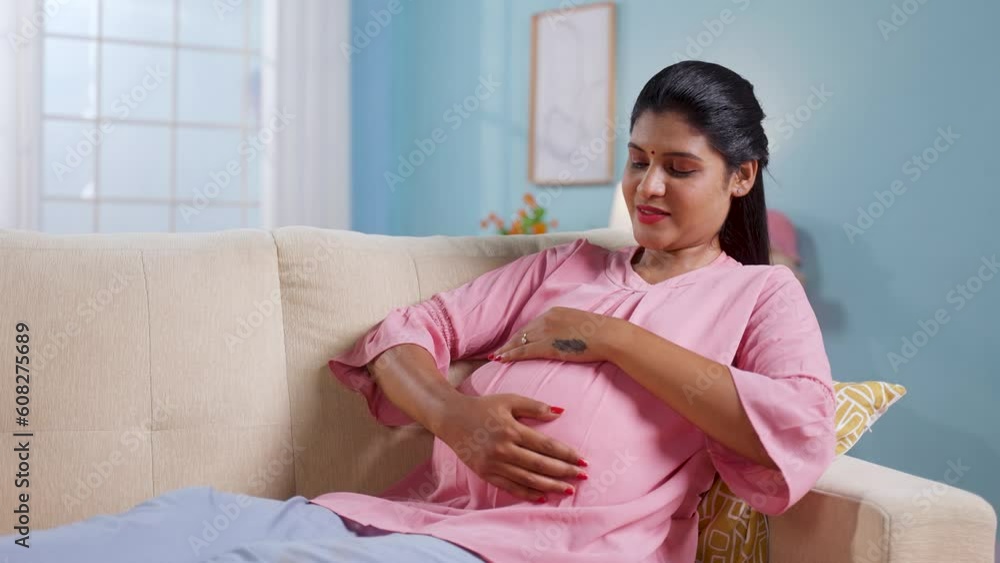 Happy indian pregnant woman feeling baby by touching tummy while sitting on sofa at home - concept of motherhood, maternity anticipation and affection Happy indian pregnant woman feeling baby by touching tummy while sitting on sofa at home - concept of motherhood, maternity anticipation and affection