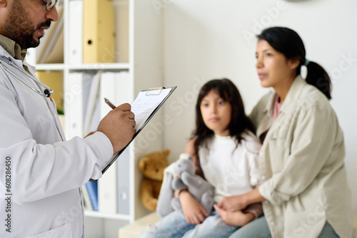 Doctor prescribing medicine to child making notes in medical card with family sitting in background