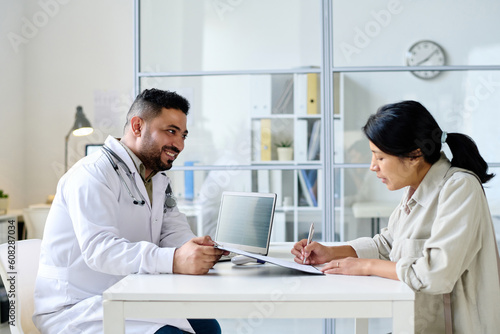 Young woman signing agreement for treatment while sitting at table with doctor in hospital