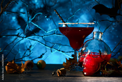 Halloween bloody cocktail margarita. Bloody red cocktail margarita and bottle form skull on scary blue dark backgrounds. Festive drink for party with candy eyes, bats, spiders. Dia de los muertos.