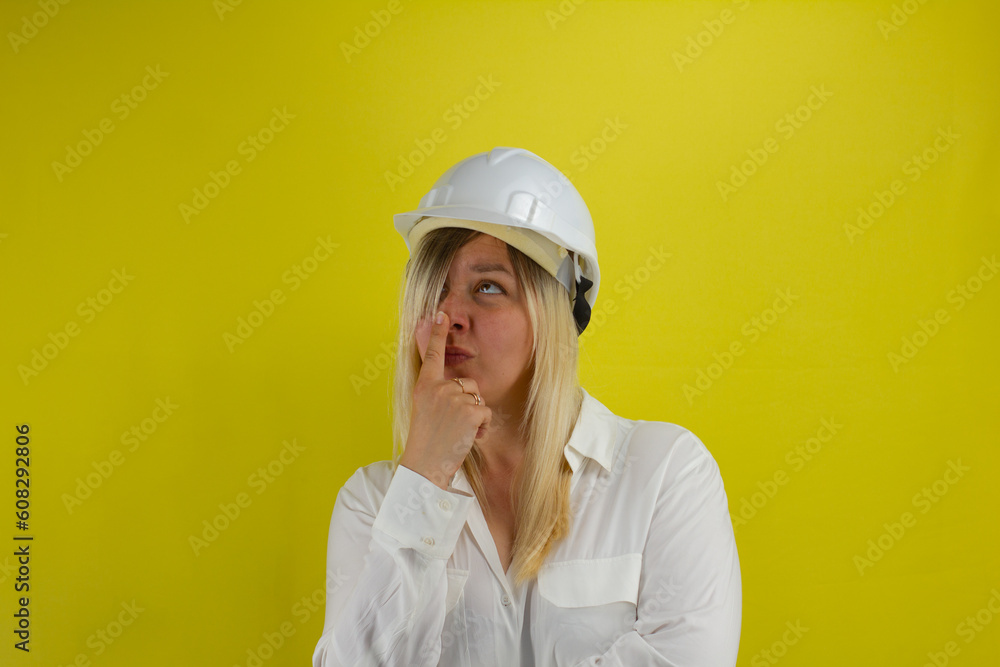 Funny female engineer in a hard hat taps her nose while thinking about solving a problem. Against a yellow background, a woman in a construction helmet looks at the ceiling in thought. Funny female engineer in a hard hat taps her nose while thinking about solving a problem. Against a yellow background, a woman in a construction helmet looks at the ceiling in thought.
