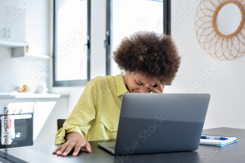 Stressed tired young Black freelance woman suffering from migraine, headache at workplace. African employee, home worker, businesswoman sitting at laptop, touching temples, holding head.. photo