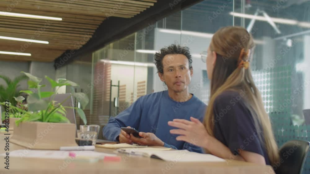 Medium shot of cheerful multi-ethnic man and woman smiling and discussing something on smartphone screen in office coworking room Medium shot of cheerful multi-ethnic man and woman smiling and discussing something on smartphone screen in office coworking room