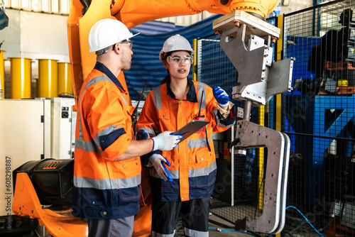 Asian female engineer and foreman caucasian in uniform wearing helmet working on machinery and quality inspection of robot arm in industrial factory.