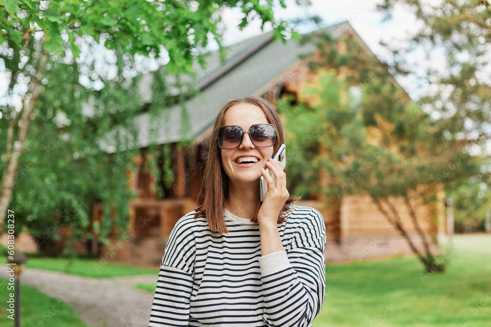 Woman wearing striped shirt and sunglasses talking on smartphone standing in park with wooden house on background laughing speaking with friends while resting in nature. Woman wearing striped shirt and sunglasses talking on smartphone standing in park with wooden house on background laughing speaking with friends while resting in nature.
