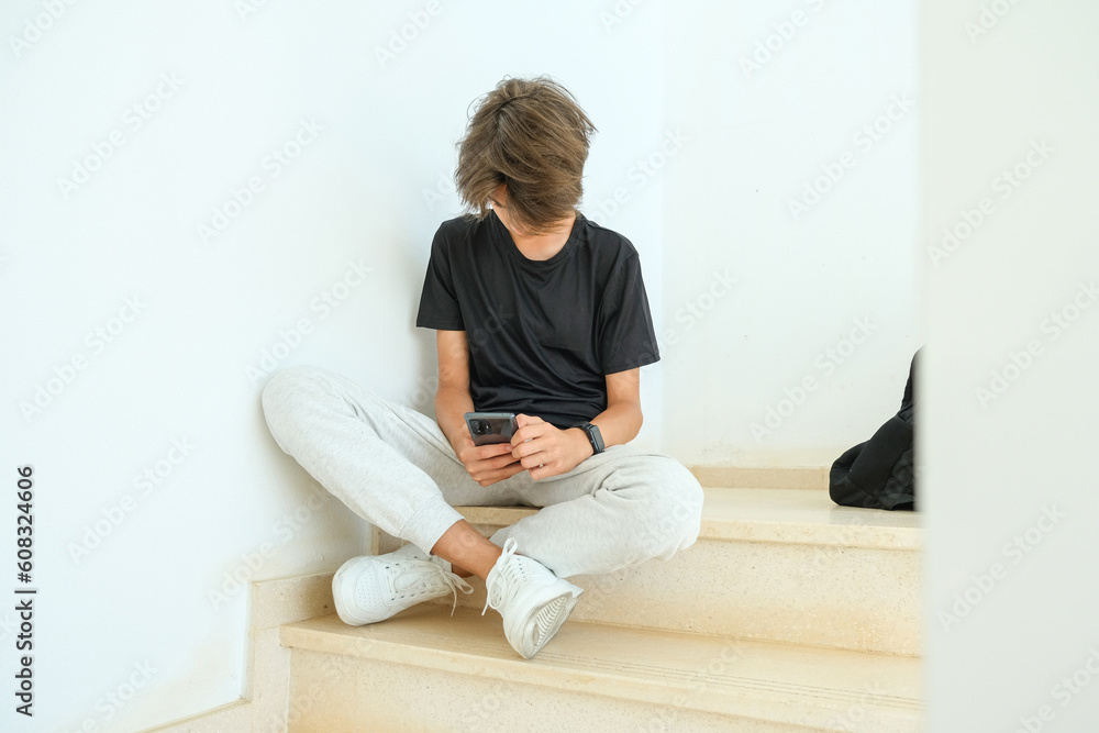 Teenage boy sitting on stairs and using smartphone. Teenager browse social media, communicate with friends, family, play games, watch videos, use apps for entertainment Teenage boy sitting on stairs and using smartphone. Teenager browse social media, communicate with friends, family, play games, watch videos, use apps for entertainment