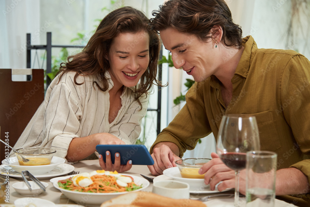 Cheerful young woman showing photo on smartphone to boyfriend when they are eating dinner at home Cheerful young woman showing photo on smartphone to boyfriend when they are eating dinner at home