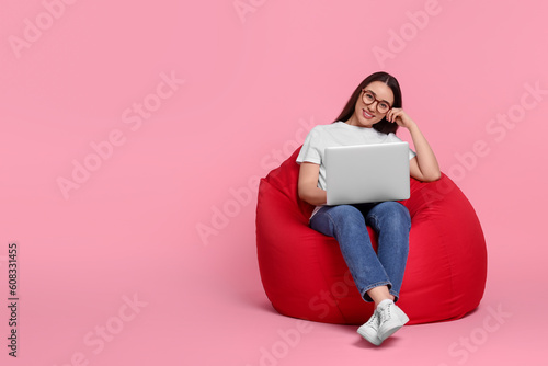 Smiling young woman with laptop sitting on beanbag chair against pink background, space for text