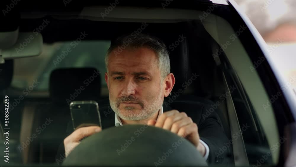 Frontal shot of a bearded man in a business suit who is sitting in a car with a phone in his hands. Man sitting at the wheel of a car and typing a message on a smartphone. Frontal shot of a bearded man in a business suit who is sitting in a car with a phone in his hands. Man sitting at the wheel of a car and typing a message on a smartphone.