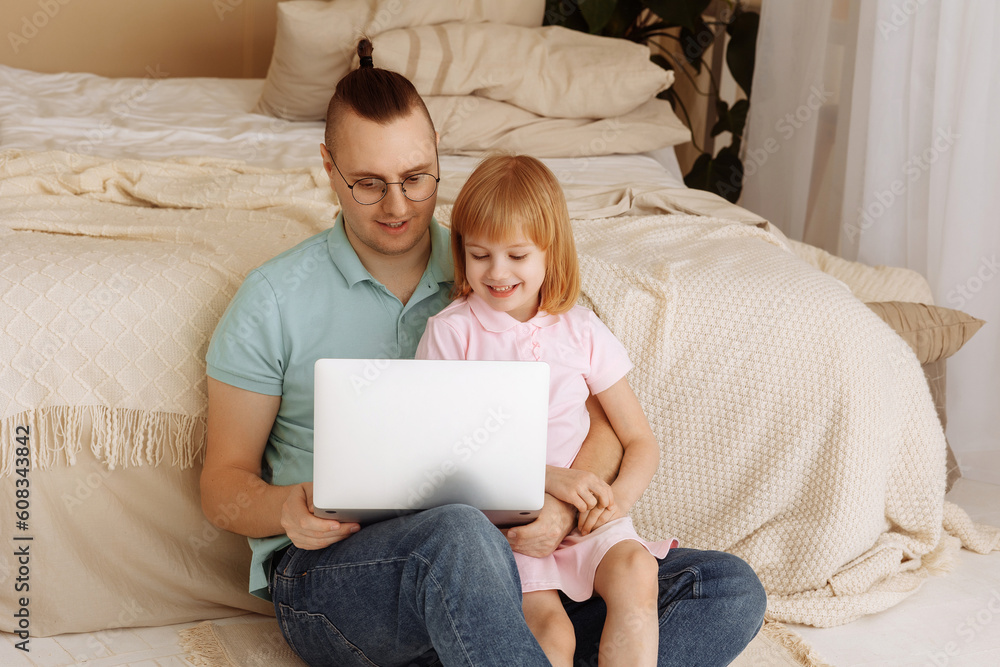 Handsome young man at home with his little cute daughter are sitting on the bed with looking into the laptop. Happy Fathers Day. Handsome young man at home with his little cute daughter are sitting on the bed with looking into the laptop. Happy Fathers Day.