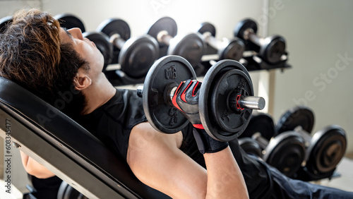 Sport man with well trained body in black sportswear doing incline dumbbell shoulder press on workout bench in fitness gym. Weight training and bodybuilding workout concept
