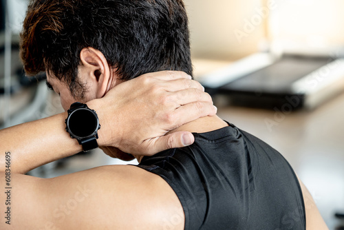 Asian sport man feeling neck pain or neckache while sitting on workout bench in fitness gym. Male athlete suffering from spine pain symptom. Sport injury concept photo