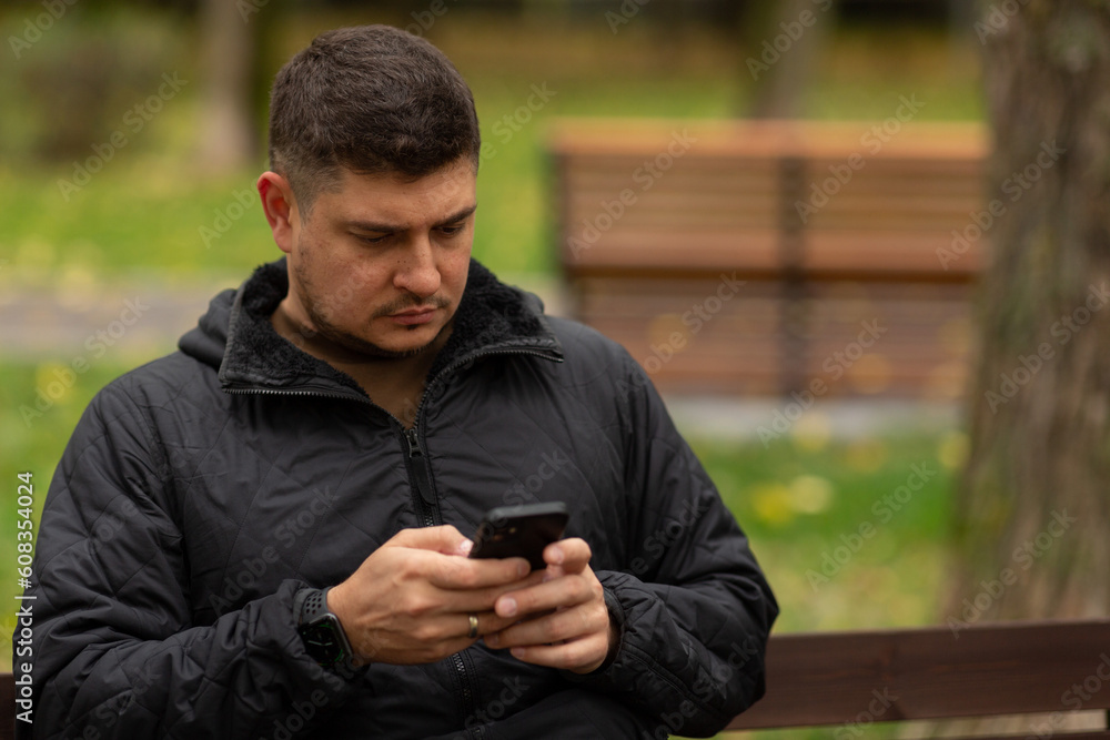 A man is sitting on a bench in the park in autumn, typing a message in his smartphone A man is sitting on a bench in the park in autumn, typing a message in his smartphone