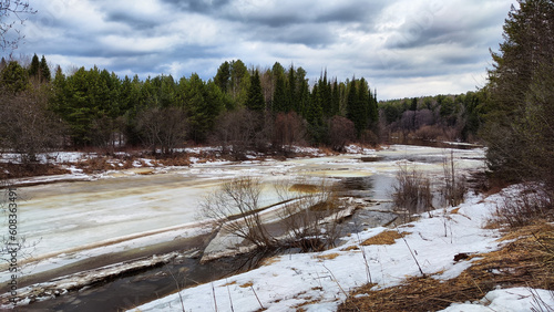 Small river with water under wet swollen ice ready for ice drift, sky with low dark clouds and forest in background in early spring afternoon. Nature landscape during a trip to the rural countryside
