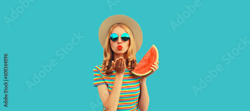 Summer portrait of beautiful young woman with fresh juicy slice of watermelon blowing her lips sends air kiss wearing straw hat on blue background