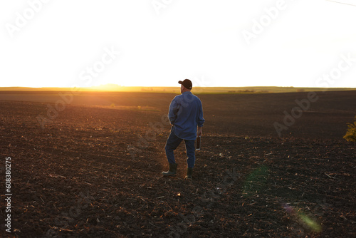Agribusiness. Back view of male farmer walking through on cultivated plowed field at sunset in spring. Owner agricultural farm is checking and examining farmland before sowing agriculture crops.