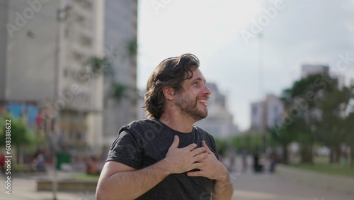 One hopeful young man putting hands in chest feeling gratitude and happiness. A joyful male caucasian person in 30s standing in city with elated emotion