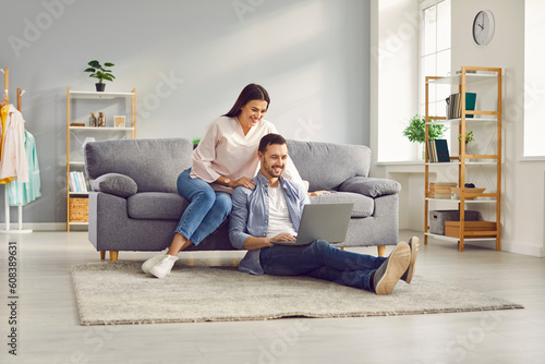 Young married couple sitting on sofa in the living room at home using tablet PC for internet and social media. Happy family resting on couch enjoying weekend watching video or talking on video call.