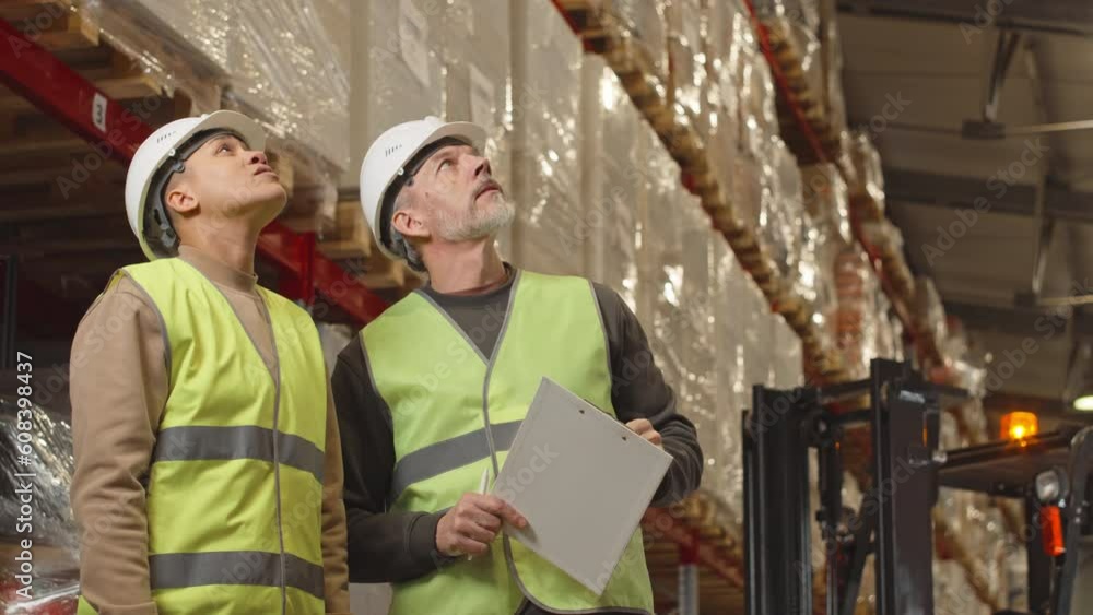 Medium shot of diverse couple of coworkers in safety uniforms looking around big warehouse and having conversation Medium shot of diverse couple of coworkers in safety uniforms looking around big warehouse and having conversation