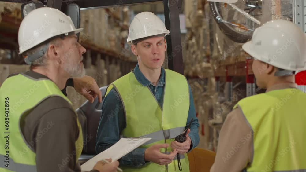 Medium shot of young Caucasian male forklift operator and his two colleagues all wearing hard hats and acid green vests having conversation during shift at warehouse Medium shot of young Caucasian male forklift operator and his two colleagues all wearing hard hats and acid green vests having conversation during shift at warehouse