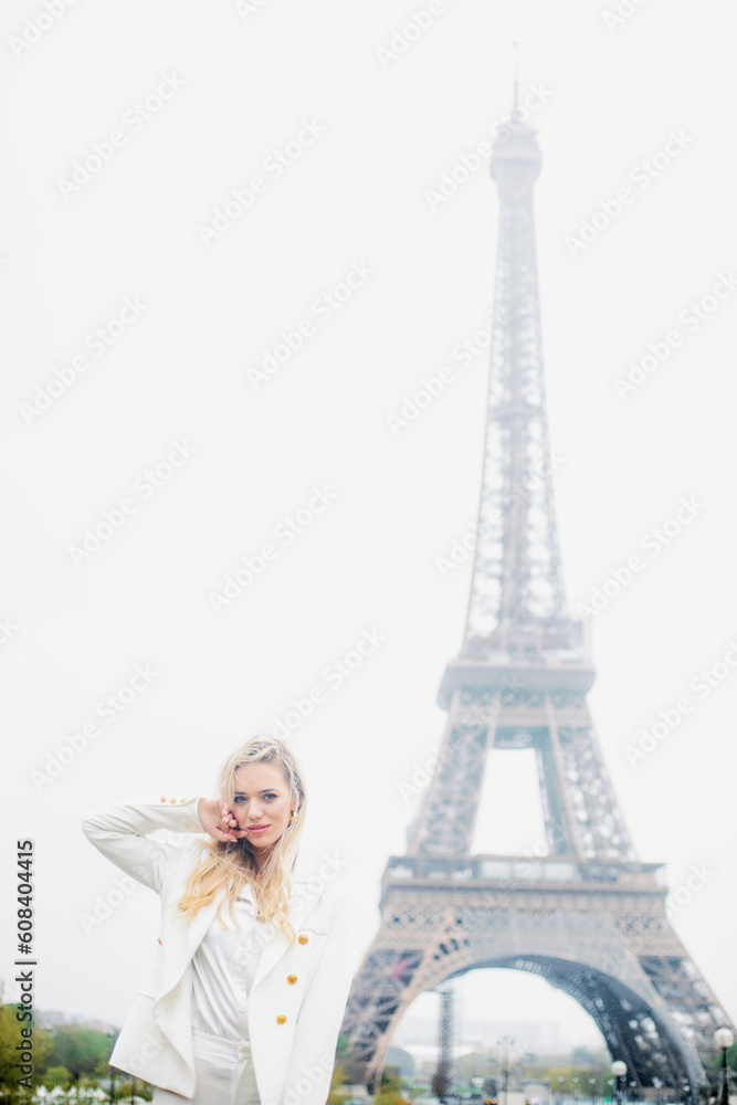Lady with flowing blond hair, in white clothes softly touching face with hand on background of famous Eiffel Tower.