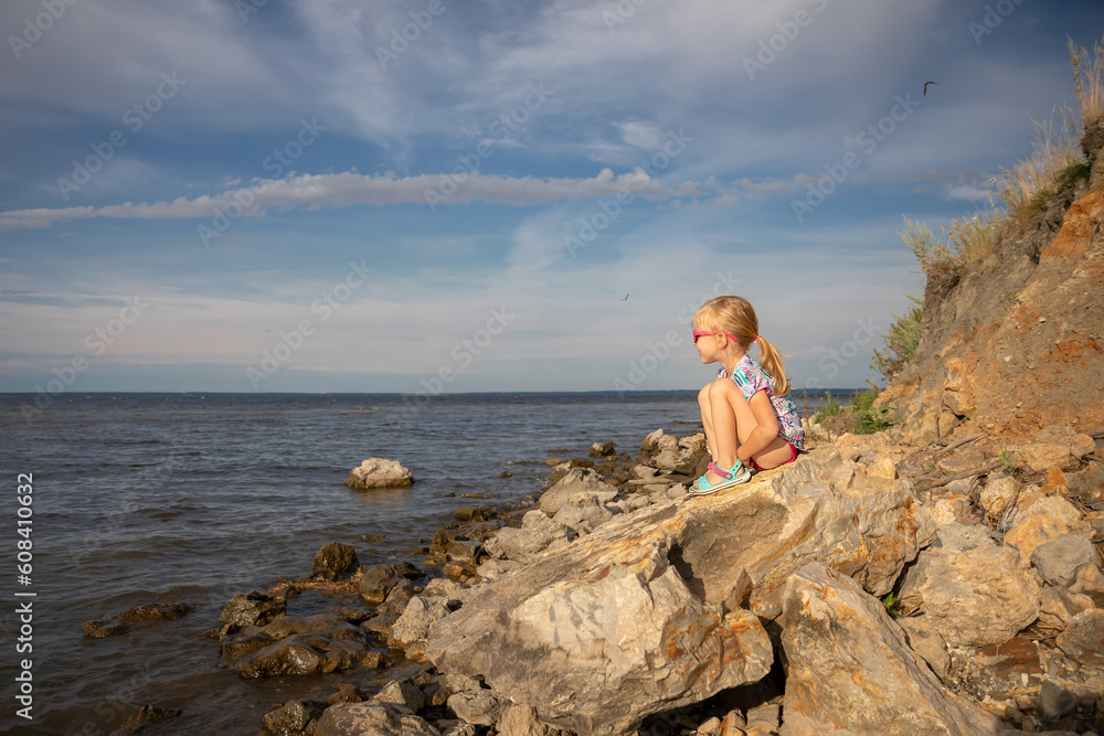 A little girl sits on a rock and looks at the sea. The concept of summer vacation, holidays and vacation at sea. copy space