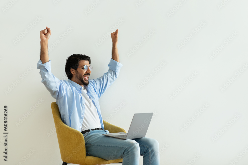 Glad excited millennial middle eastern guy in glasses rises hands up, celebrating victory, making success gesture Glad excited millennial middle eastern guy in glasses rises hands up, celebrating victory, making success gesture