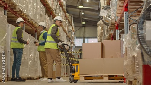 Full length shot of female warehouse worker carrying hand pallet truck loaded with parcels packed in cardboard boxes along aisle with other goods prepared for dispatching
