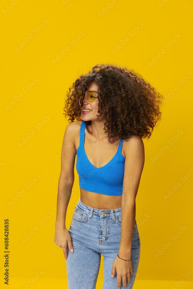 Woman with curly afro hair in a blue T-shirt on a yellow background dancing flying hair with sunglasses yellow, hand signs, look into the camera, smile with teeth and happiness, copy space Woman with curly afro hair in a blue T-shirt on a yellow background dancing flying hair with sunglasses yellow, hand signs, look into the camera, smile with teeth and happiness, copy space