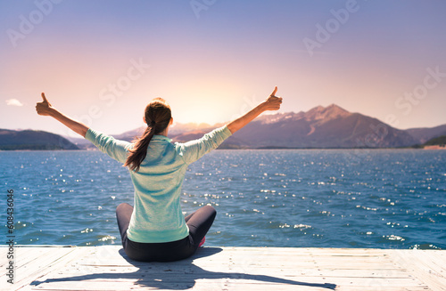Young woman facing mountain lake sunset rejoices, feeling positive, smiles looking up to the sky, enjoys life and summer, nature, happiness. 
