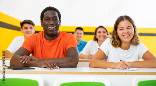 Positive african-american man and caucaisan girl studying in taxi driving school, sitting at his desk with group of people.