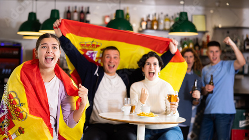 Company of excited young adult sports fans waving flag of Spain and supporting national team with beer in pub