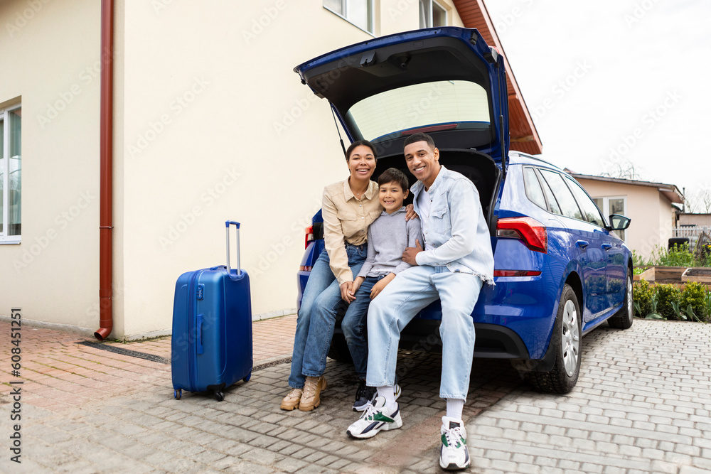 Happy young african american family of three sitting together in car trunk, ready for summer vacation trip, looking and smiling at camera near house outdoors