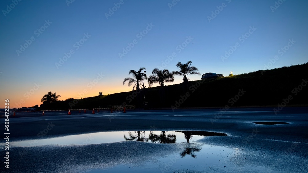 Wide view, silhouetted SUV and palm trees at top of hill with orange sunset sky in background and reflections on puddles in parking lot in foreground