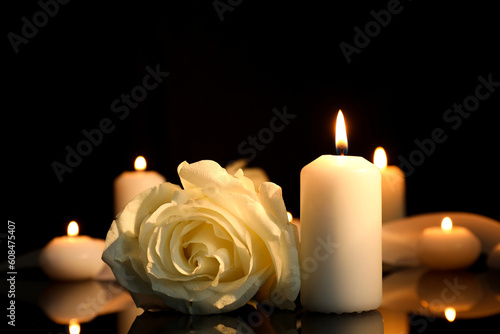 White rose and burning candles on black mirror surface in darkness  closeup with space for text. Funeral symbols