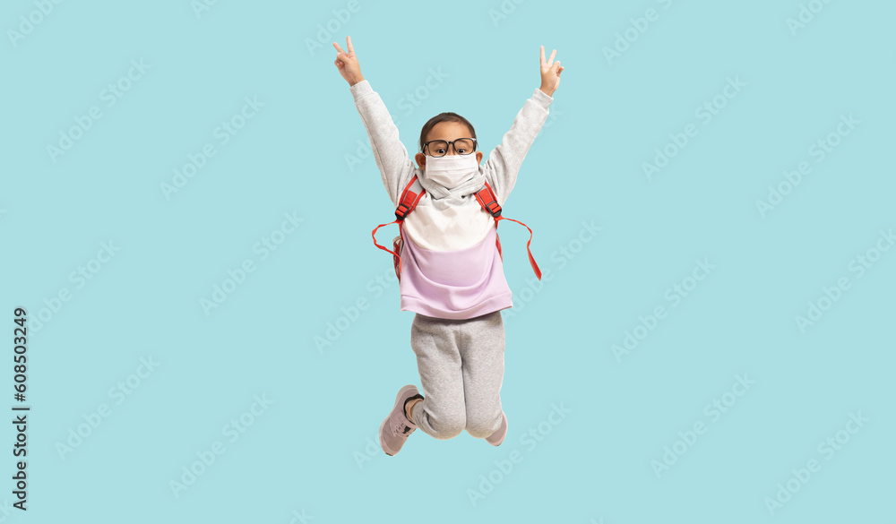 Asian little girl wearing mask showing two fingers and jumping, Full body portrait isolated on pastel plain light blue background Asian little girl wearing mask showing two fingers and jumping, Full body portrait isolated on pastel plain light blue background