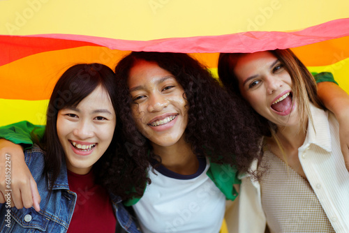 Happy cheerful LGBTQ multiethnic women posing for photography together with a rainbow flag on yellow background. 