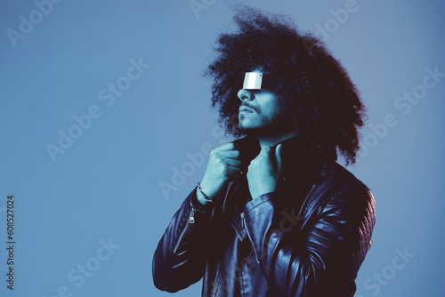 Portrait of fashion man with curly hair with stylish glasses on blue background multinational, colored light, black leather jacket trend, modern concept.