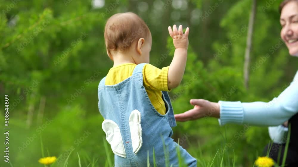 Child girl in bright overalls walks along lawn grass to mother. Positive woman stretches out hand helping toddler. Wildlife walk concept slow motion Child girl in bright overalls walks along lawn grass to mother. Positive woman stretches out hand helping toddler. Wildlife walk concept slow motion