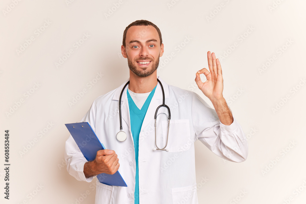 Bearded practitioner stands on blue background wearing white coat with stethoscope on his neck, holds clipboard and shows OK sign, professional people concept, copy space Bearded practitioner stands on blue background wearing white coat with stethoscope on his neck, holds clipboard and shows OK sign, professional people concept, copy space