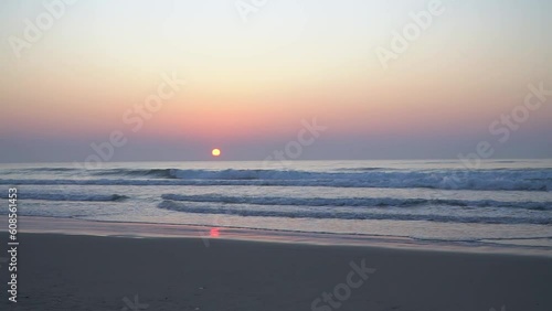 Video of sunrise landscape on sandy beach with waves and rising sun over mist, cloudless sky. And accompanied by the sound of waves and wind.