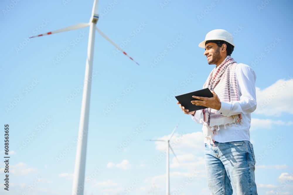 Engineer India man working at windmill farm Generating electricity clean energy. Wind turbine farm generator by alternative green energy. Asian engineer checking control electric power Engineer India man working at windmill farm Generating electricity clean energy. Wind turbine farm generator by alternative green energy. Asian engineer checking control electric power