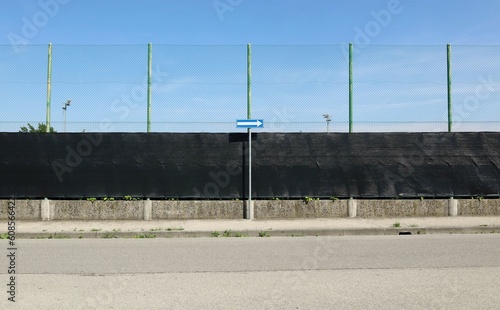 Road directional arrow sign on a concrete sidewalk. On behind a fence consisting of black tarpaulin with high wire mesh. Asphalt road in front. Background for copy space