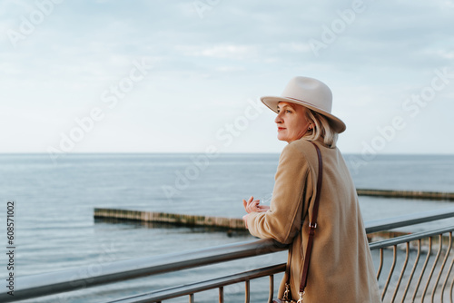 Fotografia Stylish pensive mature woman in brown round hat and coat standing on embankment and looking away at sea horizon