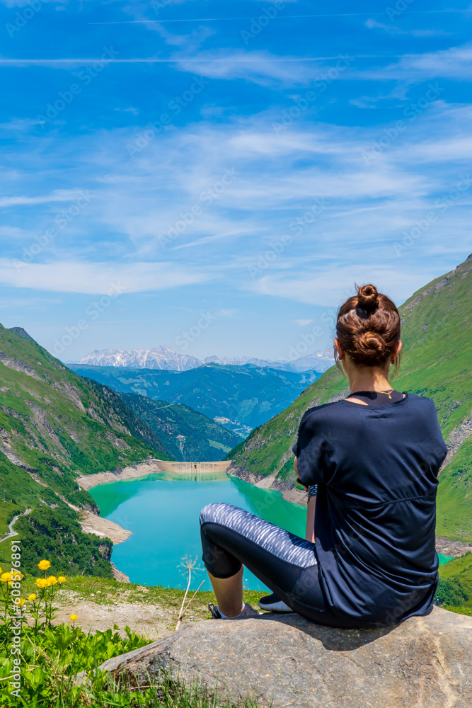 Woman sitting and looking at Kaprun water reservoirs, Kaprun dam, Austria. Stunning turquoise picturesque lake surrounded by the green Alps mountains, blue sky, and clouds. Famous hiking destination.