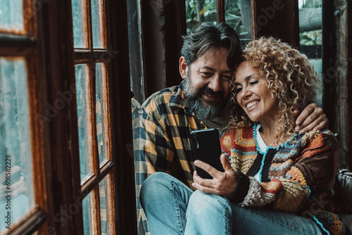 Serene romantic couple adult using phone together for video call or enjoying app smiling and sitting on the floor in home chalet. Man and woman enjoy technology and relationship in romantic atmosphere