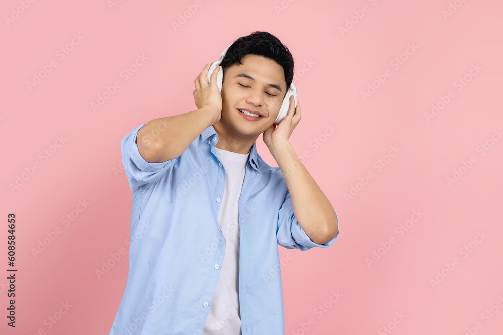 Happy asian handsome man wearing wireless headphones with closed eyes listening to favorite song standing isolated on bright pink background. for advertising