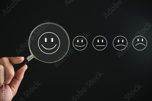 Magnifier in hand focuses on happy smiley face icon among emotions on black. Good feedback rating and positive customer review. Experience, satisfaction survey. Certificate symbolizes satisfaction.