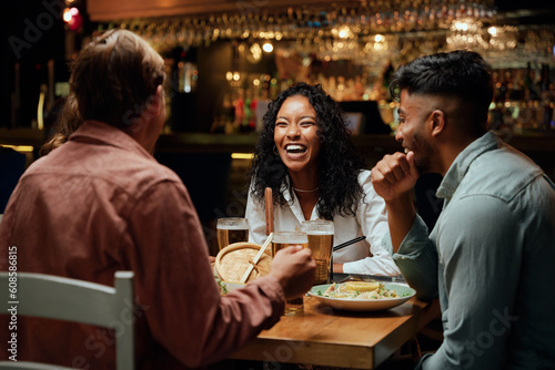 Happy young multiracial group of friends in casual clothing laughing around table with food and drinks