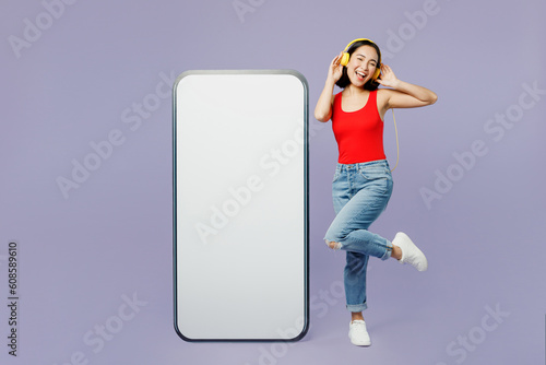 Full body young woman of Asian ethnicity wear casual clothes red tank shirt big huge blank screen mobile cell phone smartphone with area listen muisc in headphones isolated on plain purple background. photo